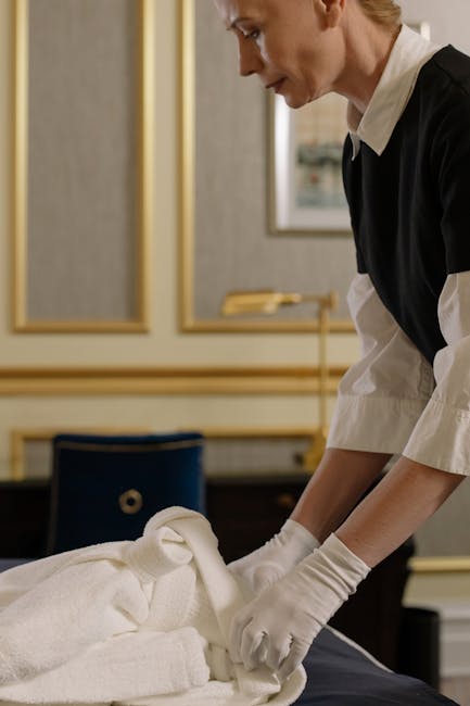A professional cleaner from Marylebone Cleaners is performing surface cleaning on a neatly folded white towel or linen on a dark wooden table in an elegant room with cream-colored walls and gold trim 