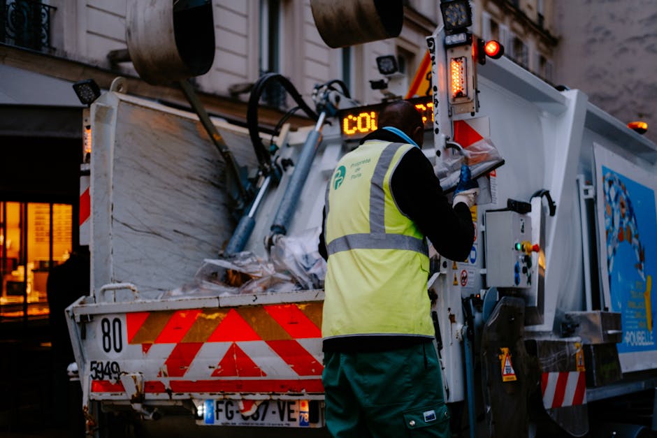 A waste collection truck parked on a residential street in Marylebone, actively engaged in disposing of bulky waste items after clearance. The truck features a white exterior with red reflective strip