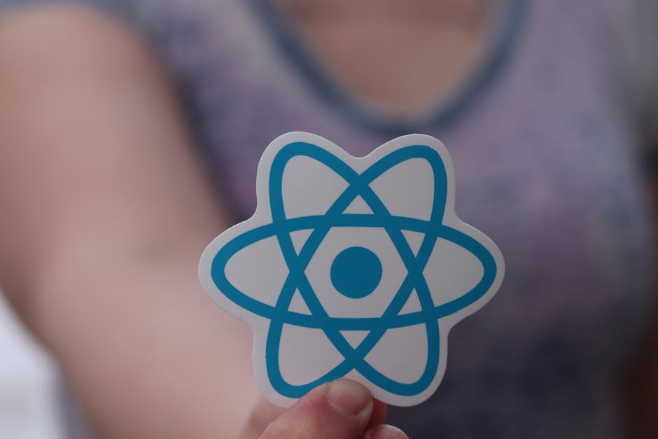 A person holding a sticker with a blue atomic symbol in front of their chest. The background shows a blurred individual wearing a grey top, with soft lighting highlighting the sticker, which is positi
