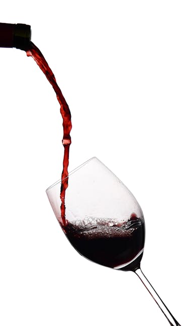 The image shows a close-up of a wine glass on a white surface, positioned on a reflective table, with a stream of red wine being poured from a bottle into the glass. The wine glass is transparent with