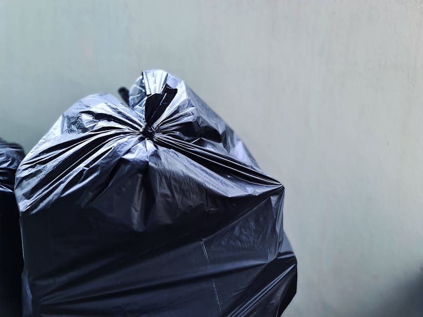 A large heap of mixed household and commercial waste, including plastic bottles, cartons, containers, and packaging, piled against a dark wooden fence. The waste materials are scattered on a concrete
